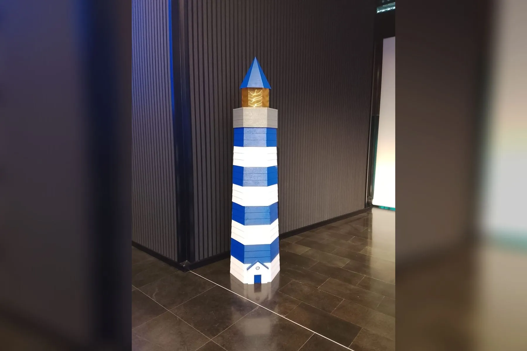 Faro decorativo de 2,5 metros impreso en 3D, a franjas azules y blancas y con la linterna iluminada, en el hall de un evento en Tenerife